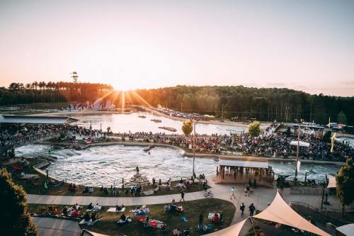 A vibrant outdoor event at sunset, with a crowd gathered around a water park featuring a wave pool and activities.