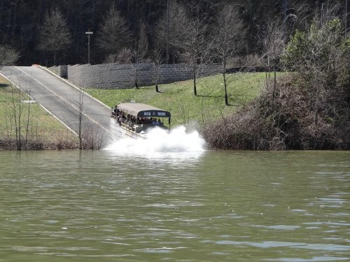 A boat splashes into a lake from a ramp, surrounded by trees and grassy banks.