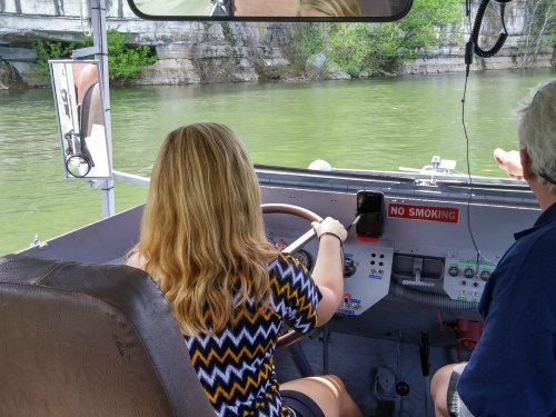 A person with long hair steers a boat on a green river, with another person seated beside them.