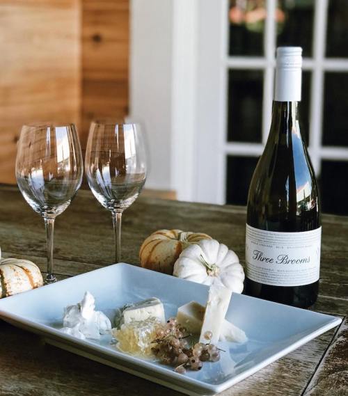 A bottle of wine with two glasses, a cheese platter, and decorative pumpkins on a wooden table.
