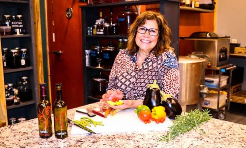 A smiling woman stands behind a kitchen counter with fresh vegetables and bottles of oil, ready to cook.