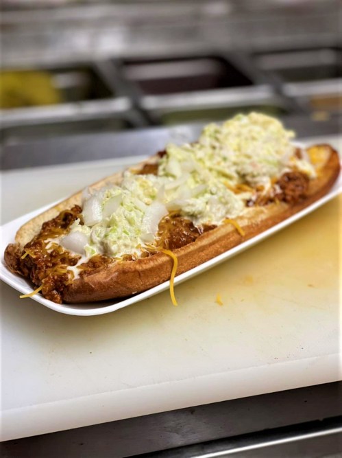 A long sandwich topped with shredded lettuce, diced onions, cheese, and a savory meat filling on a white plate.
