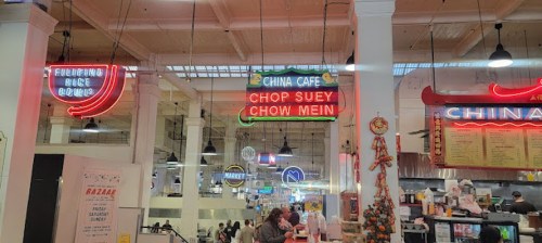 Neon signs for "China Cafe" and "Chop Suey Chow Mein" in a bustling food market with various stalls.