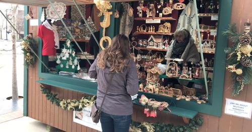 A woman browses handmade crafts at a festive market stall, with various wooden decorations displayed.