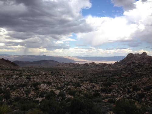 A vast desert landscape with rocky formations under a cloudy sky, showcasing distant mountains and greenery.