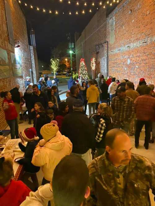 A lively outdoor gathering at night, with people enjoying festivities, holiday lights, and decorated trees in a brick alley.