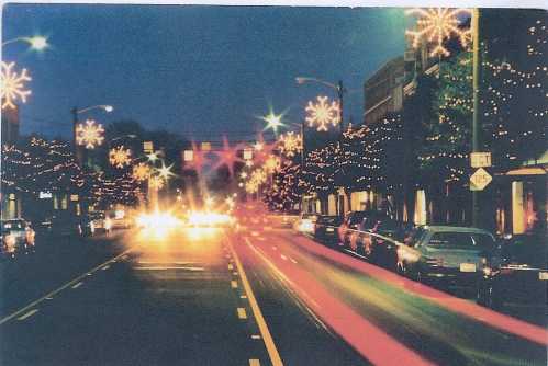 A city street at night adorned with festive lights, featuring blurred car headlights and decorated trees along the road.