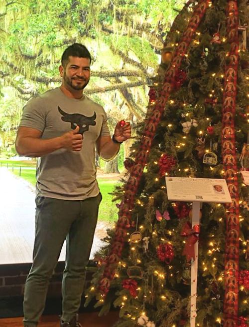 A man stands next to a decorated Christmas tree, smiling and giving a thumbs up, with a scenic background.