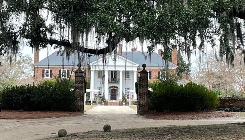 A grand white mansion with a large front porch, framed by trees and Spanish moss, set in a lush garden.