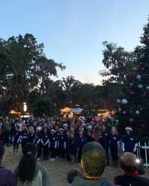 A large group of children in festive attire singing in front of a decorated Christmas tree at an outdoor event.