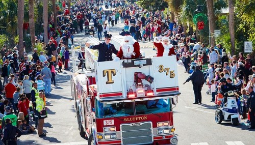 A festive parade scene with a fire truck carrying Santa and a police officer, surrounded by a large crowd.