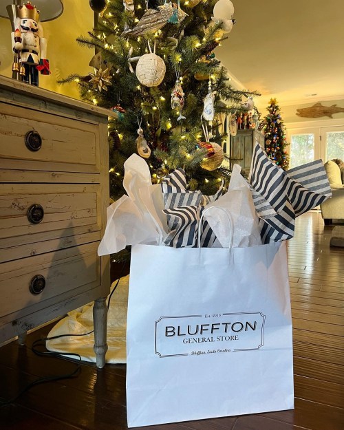 A festive scene featuring a Christmas tree and a gift bag from Bluffton General Store, filled with wrapped presents.