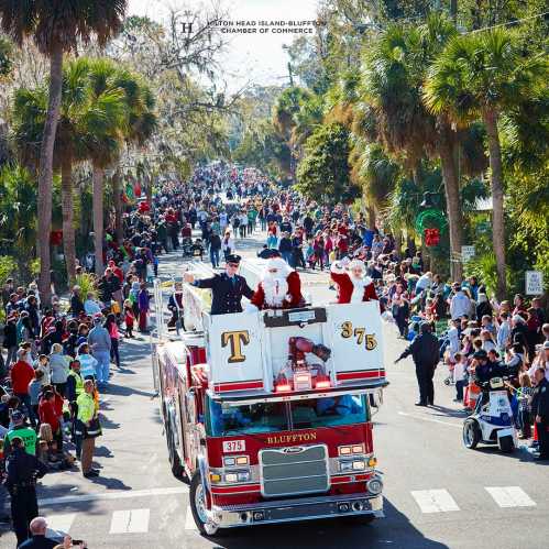 A festive parade scene with a fire truck carrying two Santa figures, surrounded by a large crowd and palm trees.