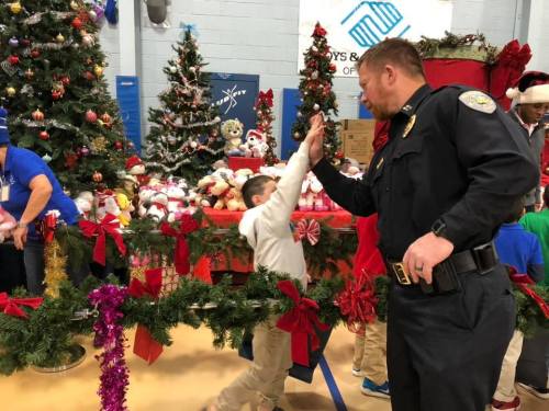 A child high-fives a police officer in a festive setting with Christmas trees and decorations in the background.