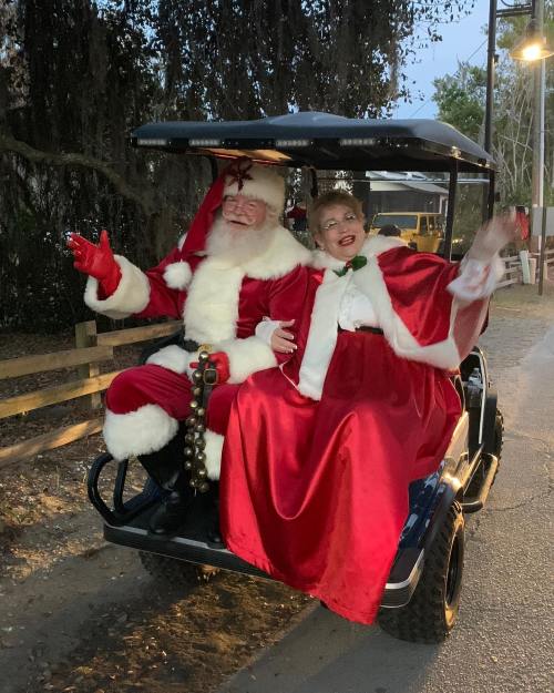 Santa Claus and a woman in festive attire wave from a golf cart, surrounded by trees and evening lights.