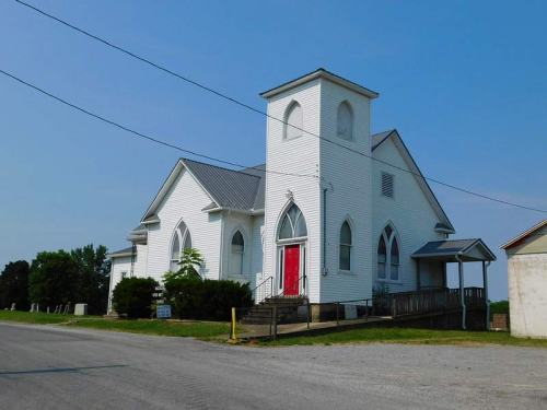 A white church with a red door and tall windows, set against a clear blue sky and surrounded by greenery.