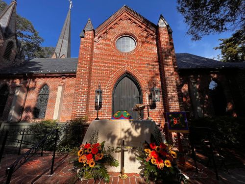 A brick church exterior with a dark door, surrounded by colorful flowers and a small altar in front.