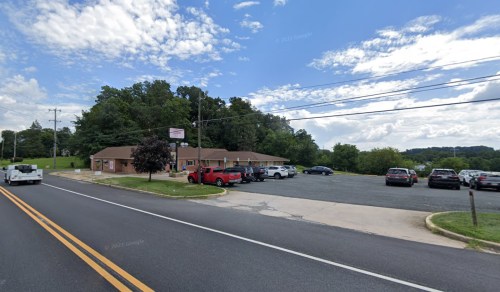 A roadside view of a small building with a parking lot, surrounded by trees and under a partly cloudy sky.