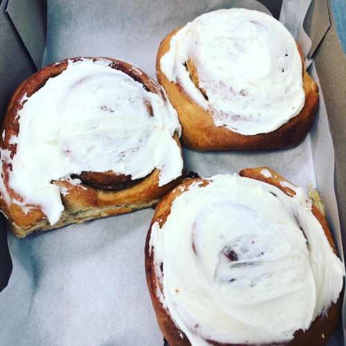 Three frosted cinnamon rolls in a cardboard box, resting on a sheet of parchment paper.