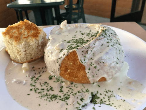 A bread bowl filled with creamy soup, garnished with herbs, alongside a piece of bread.
