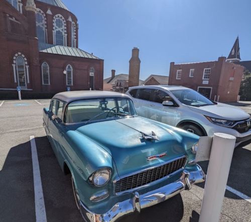 A vintage turquoise car parked next to a modern white SUV in a sunny lot with a church in the background.