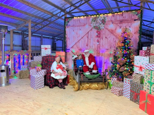 A festive scene with Santa, two children, and a decorated Christmas tree surrounded by wrapped gifts in a barn setting.