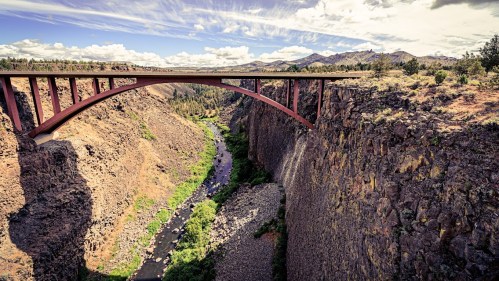 A modern bridge spans a deep canyon, with a river flowing below and mountains in the background under a cloudy sky.