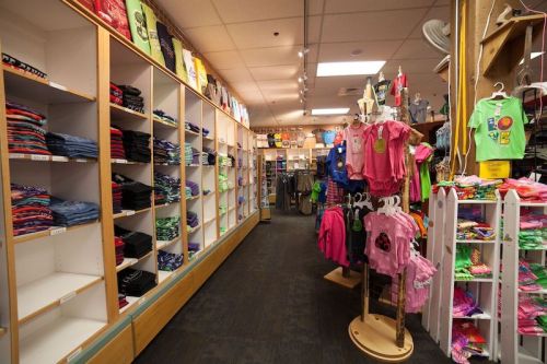 A retail store aisle with shelves of folded clothes, colorful t-shirts, and accessories displayed on racks.
