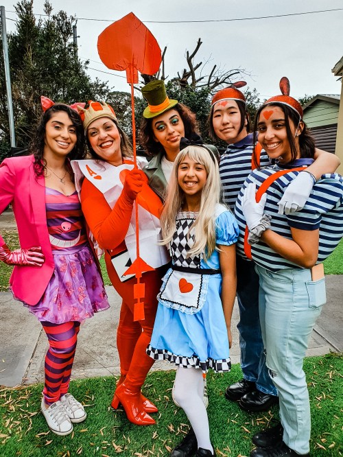 A group of six people in colorful costumes, including Alice and the Queen of Hearts, posing together outdoors.