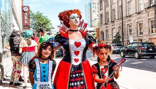 A woman dressed as the Queen of Hearts stands with two girls in costume, surrounded by a lively street scene.