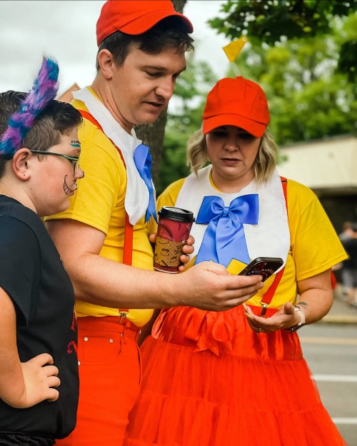 A group of three people in colorful costumes, looking at a phone, with one holding a coffee cup.