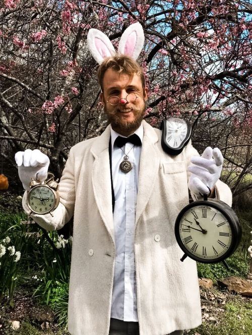 A person in a white coat and bunny ears holds three clocks in a garden with blooming flowers.