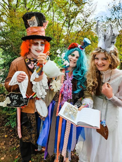 Three people dressed in colorful, whimsical costumes inspired by a tea party, surrounded by greenery.