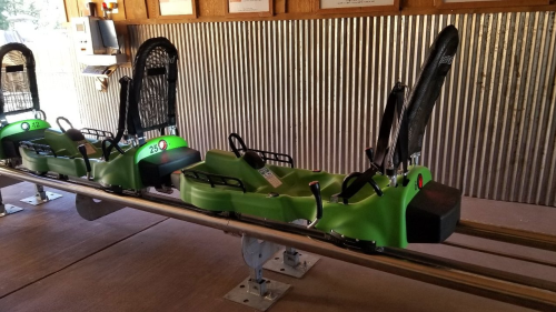 A green roller coaster cart on a metal track, positioned indoors with a rustic backdrop.