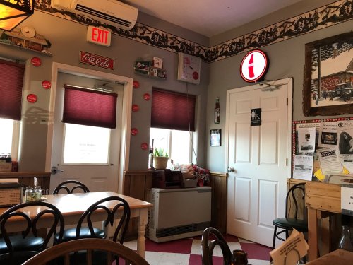Cozy diner interior with red and white decor, tables, and a Coca-Cola sign on the wall. Windows with blinds are visible.