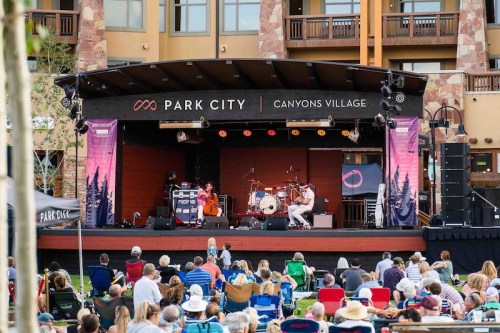 A live music performance on stage at Canyons Village, with a crowd seated on the grass enjoying the show.