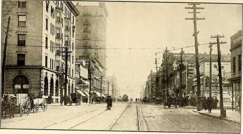 Historic street scene with horse-drawn carriages, early 20th-century buildings, and power lines.