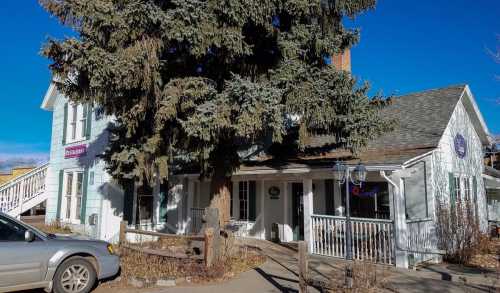 A charming two-story house with a large tree in front, featuring a porch and a sign, set against a clear blue sky.