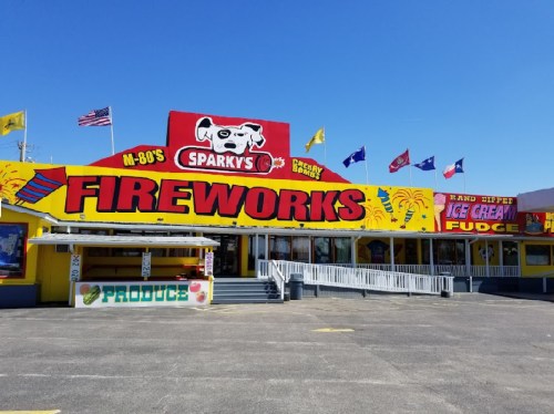 Brightly colored storefront of Sparky's Fireworks, featuring flags and signs for ice cream and produce. Clear blue sky above.