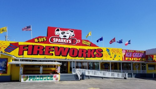 Brightly colored storefront of Sparky's Fireworks, featuring flags and signs for ice cream and fudge, under a clear blue sky.