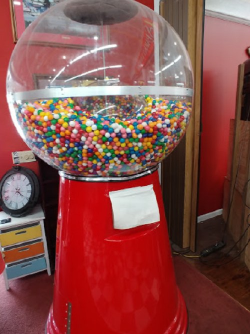 A large gumball machine filled with colorful gumballs, set against a red wall in a cozy interior.