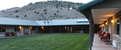 A courtyard view of a motel with a grassy area, surrounded by mountains and outdoor seating along the building.
