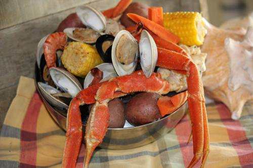 A bowl filled with various seafood, including crab, clams, corn, and potatoes, arranged on a checkered cloth.