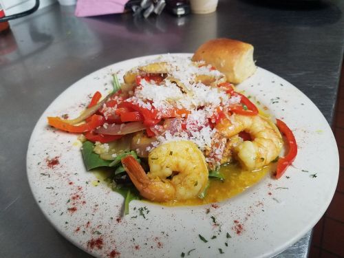 A plate of shrimp with colorful peppers, onions, and cheese, served on a bed of greens with a roll on the side.