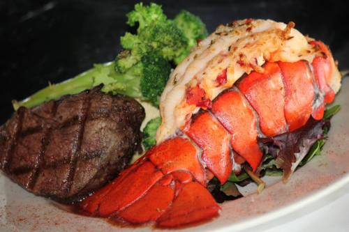 A plate featuring a grilled lobster tail, steak, and steamed broccoli on a bed of mixed greens.