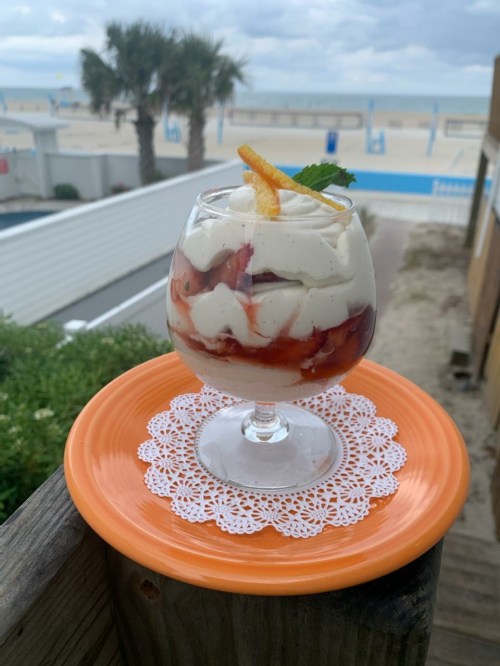A glass dessert with layers of cream and fruit, garnished with a mint leaf and orange peel, on an orange plate by the beach.
