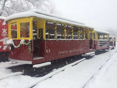 A colorful vintage train car decorated for the holidays, covered in snow, with a wintery landscape in the background.