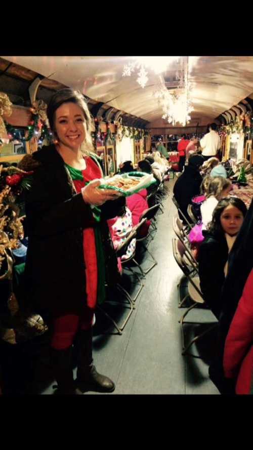 A woman in festive attire holds a tray of cookies on a decorated train filled with people enjoying the holiday spirit.