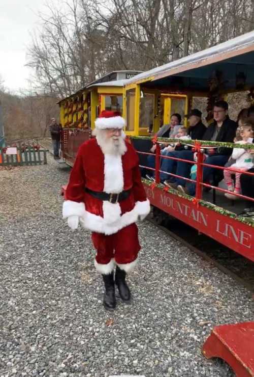 Santa Claus walks beside a colorful train, with families seated inside, surrounded by trees and gravel.