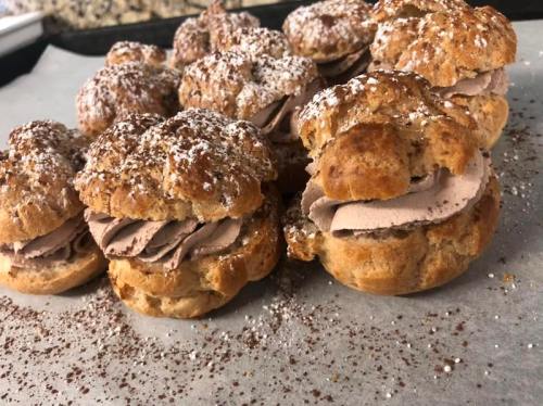 A close-up of cream puffs filled with chocolate cream, dusted with powdered sugar on a baking sheet.